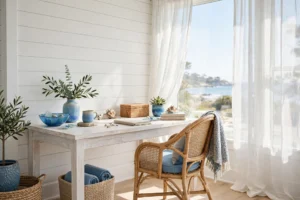 A bright coastal home office with a whitewashed desk, rattan chair, shiplap wall, and large sunlit window.